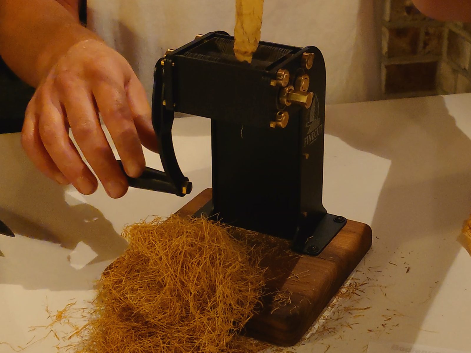 Eine Hand an der Kurbel des FINECUT PRO, Messingschrauben am Stahlrahmen, Sockel aus Edelholz, davor frischer Feinschnitt